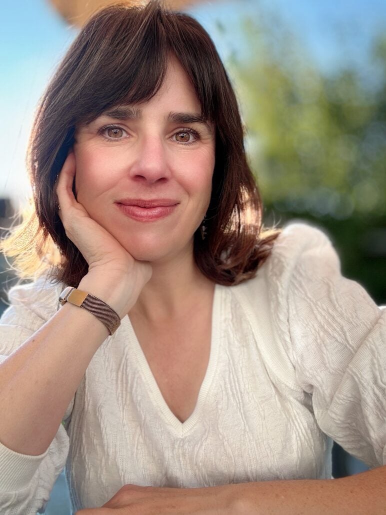 Laura Durham rests her face on one hand and smiles at the camera while seated outdoors in a white sweater.