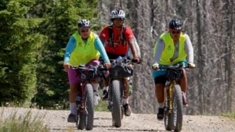 Three mountain bikers cycle along a gravel trail near the woods.