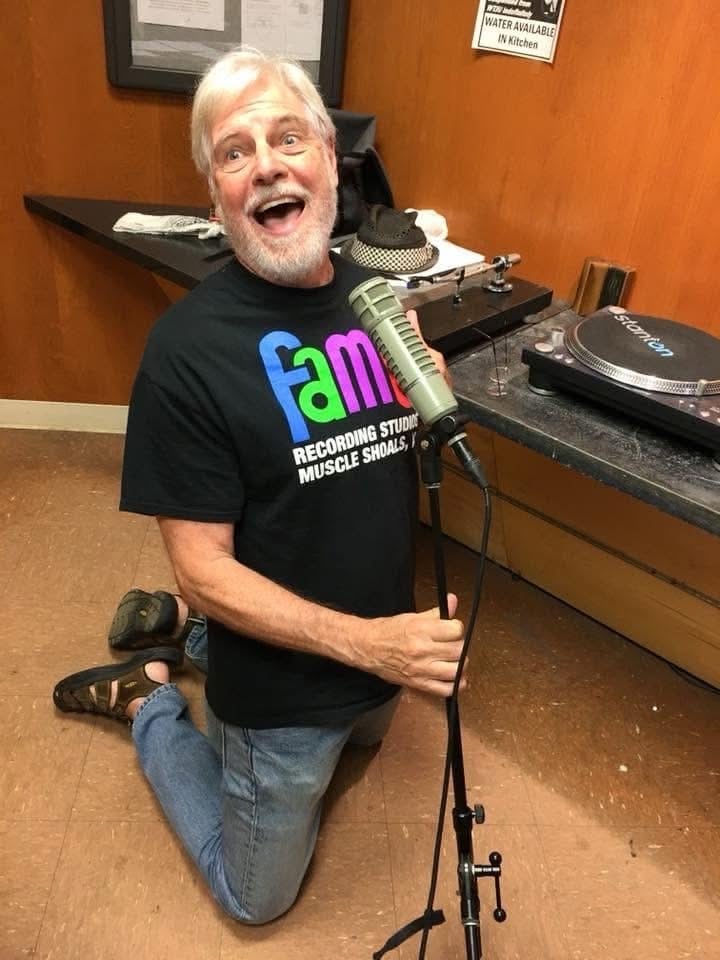 Dave Rogers kneels enthusiastically at a microphone in a radio studio, with a turntable and audio equipment behind him.