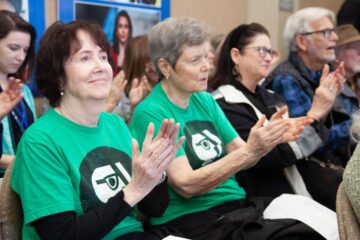 Supporters watch live coverage of an Arkansas TV commission meeting held March 12. Audience members applaud, with two people in the front row wearing matching green T-shirts featuring a PBS-style logo.