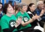 Supporters watch live coverage of an Arkansas TV commission meeting held March 12. Audience members applaud, with two people in the front row wearing matching green T-shirts featuring a PBS-style logo.