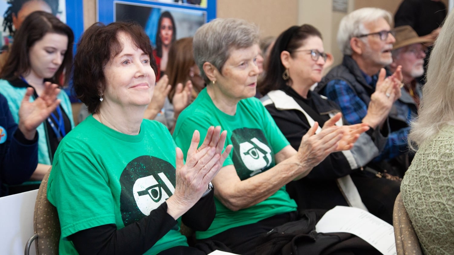 Supporters watch live coverage of an Arkansas TV commission meeting held March 12. Audience members applaud, with two people in the front row wearing matching green T-shirts featuring a PBS-style logo.