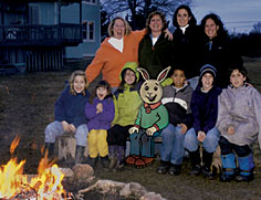 Four moms, six kids and a cartoon rabbit pose around a backyard campfire.