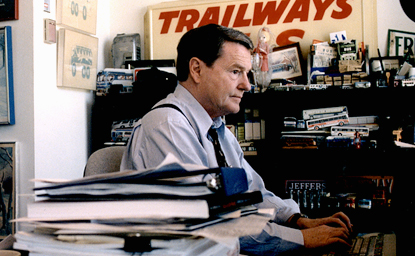 Jim Lehrer working, Trailways bus sign on wall behind him, stacks of reports on his desk